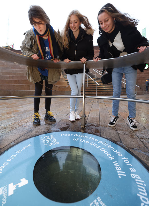 3 young people looking over a railing at a round glass panel in the pavement