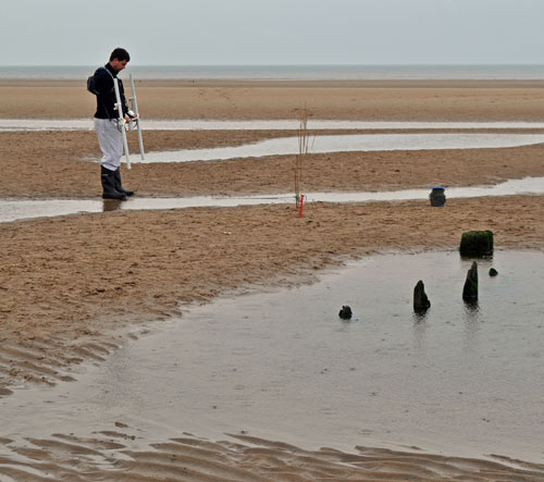 Someone using technical equipment to survey a partly-exposed shipwreck on a beach