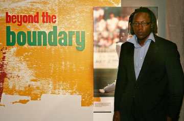 A man wearing glasss stands in front of a yellow sign that says 'Beyond the Boundary'