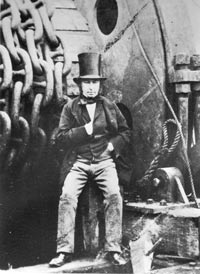 Black and white photo of a man in a top hat and waistcoat leaning against a huge wheel of chains