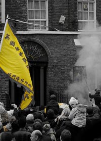 Black and white photograph of a crowd and smoking firecrackers, with flag and other details picked out in yellow