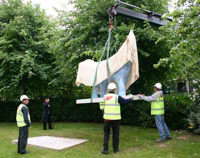 Photo showing a large lamb-shaped sculpture being whinched into place on a lawn by a large crane. People in hard hats are supervising.