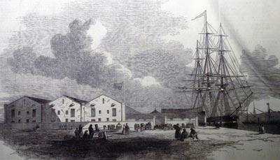 Etching of a ship docked next to warehouses.