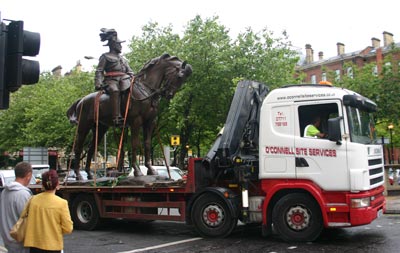 Monument of man on horse strapped to the back of a flat bed lorry, watched by pedestrians