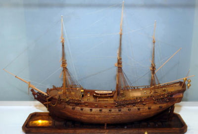 Colour photo of a wooden ship model. It has 3 large masts and a small boat on the deck.