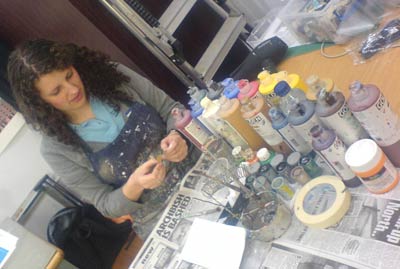 a dark haired young woman painting on a table surrounded by paint pots