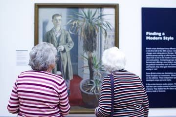 Two women look at a painting on the wall of a gallery