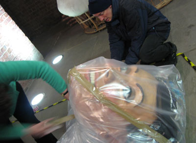 two people wrapping a giant model head in plastic sheeting