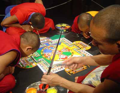 four men in red robes bending over a colourful pattern