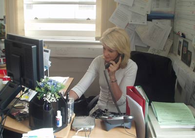 Photo of a blond woman sitting at a computer screen and speaking on a telephone