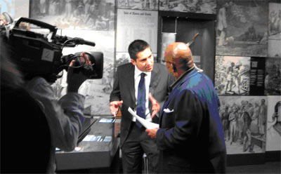 two men talking on a museum gallery while being filmed by a man with a large camera