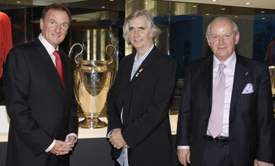 Three men in suits standing in front of a glass case containing a large silver trophy