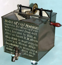 colour photograph of a black box with a wooden handle protruding from the top, a tap at the front and writing across the front of the box.
