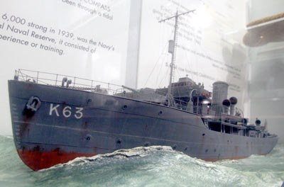 Photo of a model of a grey ship at sea. It has a red hull and the number K63 on its side