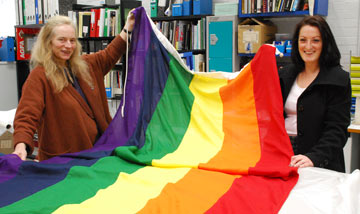 two women holding up a large flag with broad stripes in rainbow colours