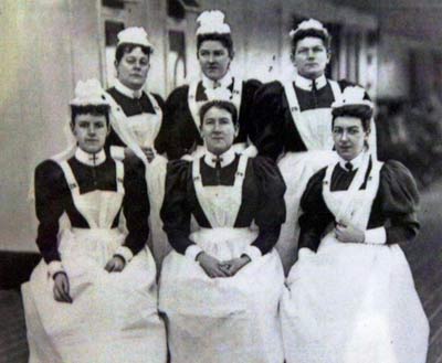Black and white photo of six women in white aprons and hats posing on the deck of a ship