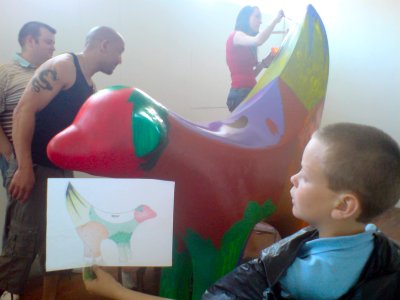 People painting a large lamb-like sculpture while a boy holds up a drawing of what it should look like