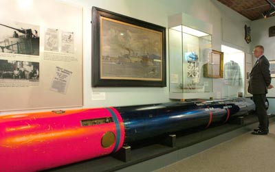 man in a museum standing next to a long torpedo, text panels and cases