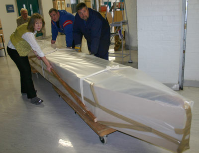 four people moving a long thin boat wrapped in plastic sheeting on trolleys down a corridor