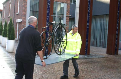 Two men carefully carry a very old bicycle into a building