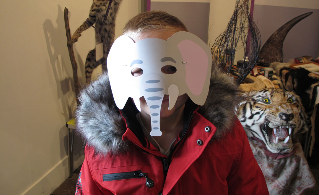 boy wearing paper elephant mask by a display including taxidermy animals and a tiger skin rug