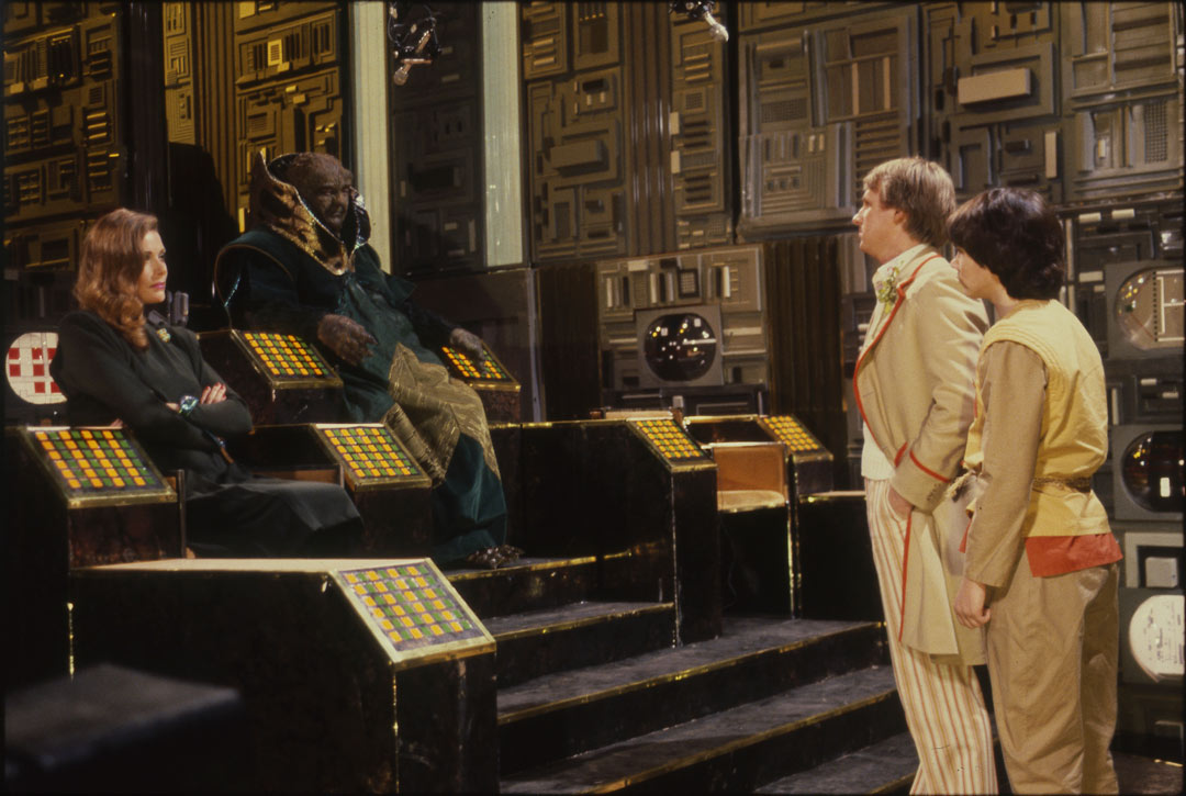 Peter Davison's Doctor and companion facing aliens on a throne