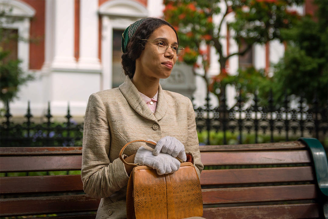 Actor in costume as Rosa Parks, a smartly dressed young Black woman, sitting on a wooden bench