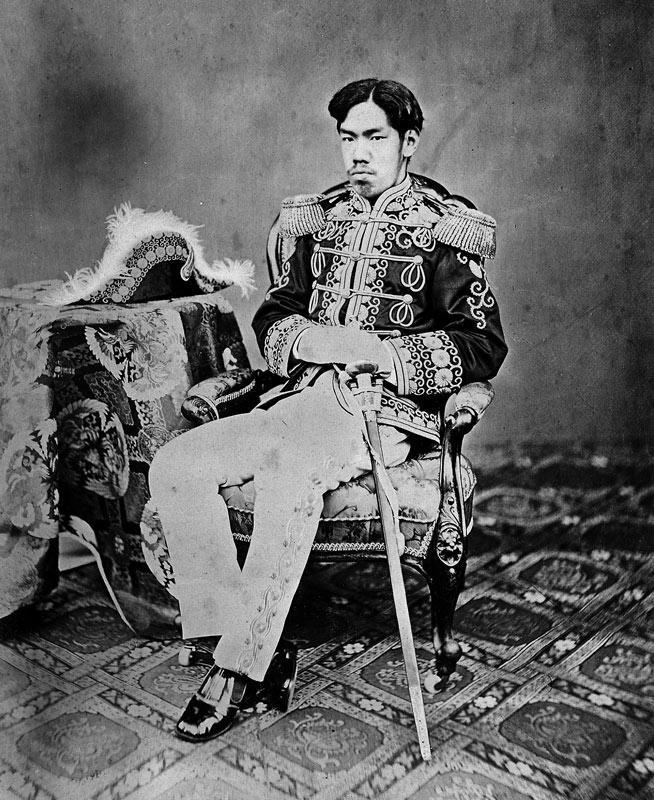 Emperor dressed in decorative braided military jacket and trousers, sitting with a plumed hat next to him on a table with a sword at his side