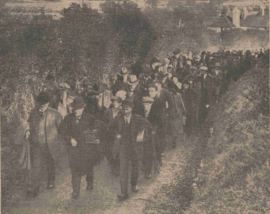 Funeral procession of many people walking up a hill, led by a man carrying a small casket