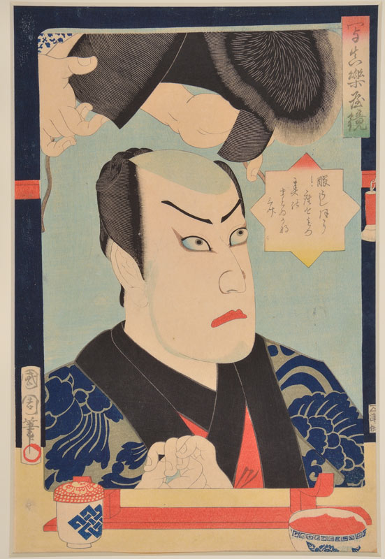 face of an actor in traditional Japanese dress, reflected in a mirror. Hands are lowering a wig into his head