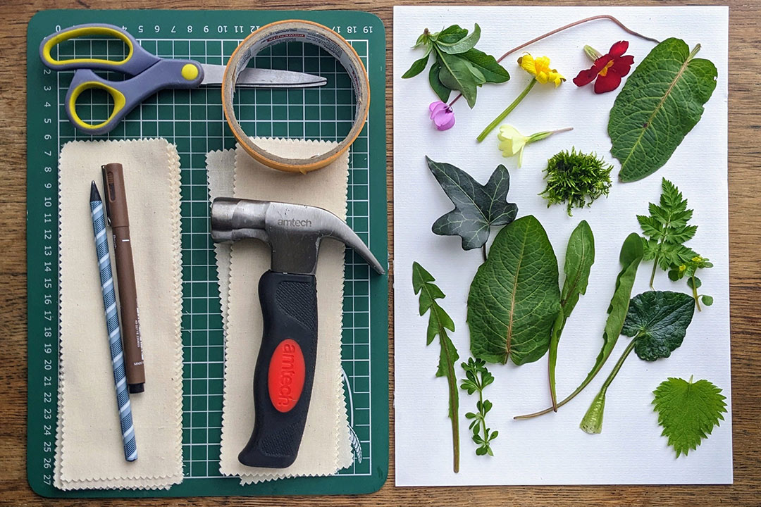 scissors, hammer, roll of masking tape, pen, pencil and fabric strips on a cutting board next to leaves and flowers on white paper