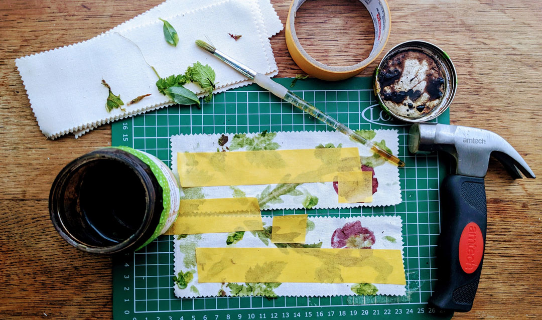 flower prints on a cutting board with masking tape over some areas, with a jar of ink, paintbrush and hammer around them
